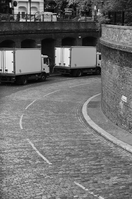 A narrow urban street scene showing a row of parked cars along the right side of the road, with a black van and a white delivery truck further ahead. The street is lined with older brick and stone bui