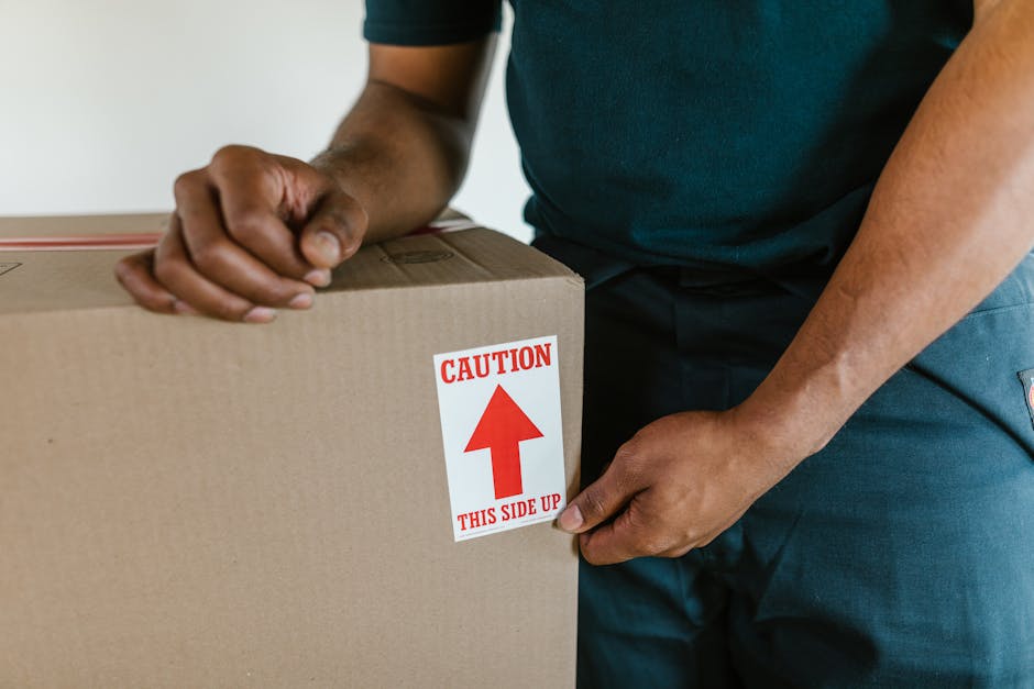 A person wearing a dark shirt and blue jeans is gripping the corner of a large cardboard box during a home relocation process, with a red and white caution label displaying an upward arrow and the wor