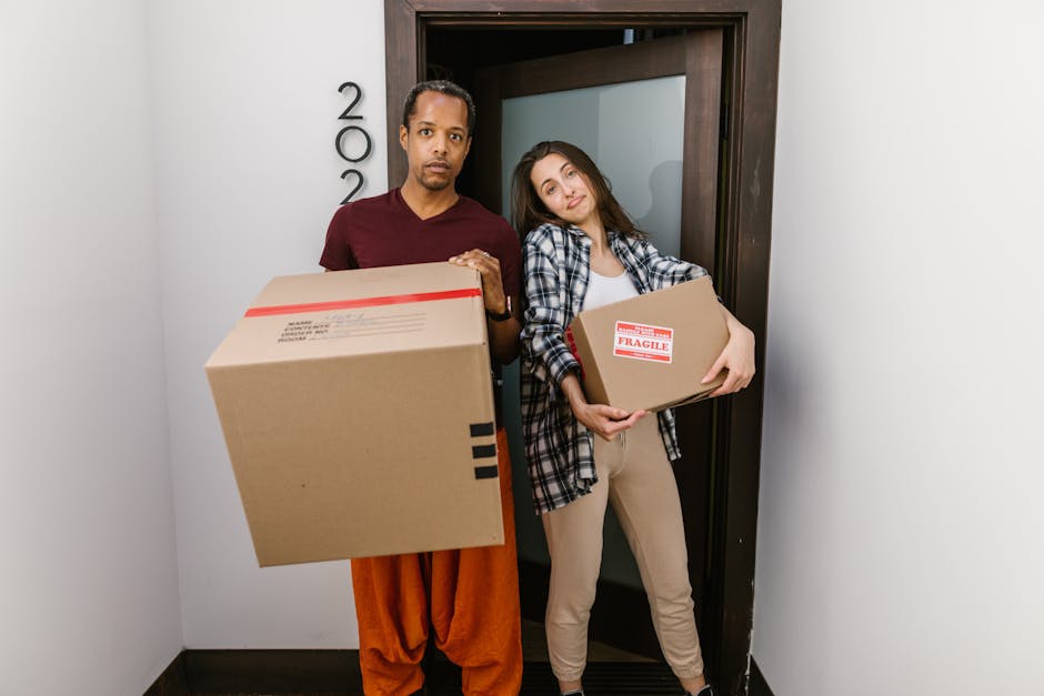 A person wearing a dark shirt and blue jeans is gripping the corner of a large cardboard box during a home relocation process, with a red and white caution label displaying an upward arrow and the wor
