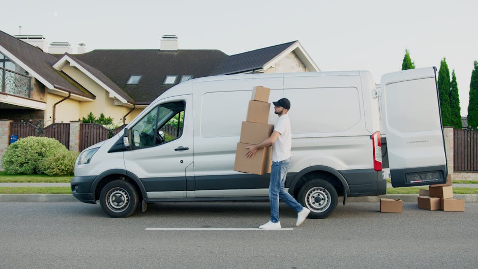 Two movers from Man and Van Finsbury Park are engaged in a home relocation process during daytime, lifting and loading cardboard boxes onto a trolley in a residential driveway. One worker, dressed in 