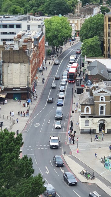 An aerial view of a city street during daytime, showing multiple vehicles including cars, vans, and a double-decker bus moving along a curved road lined with trees and buildings. On the left side of the street, pedestrians walk on the pavement, some waiting at the corner near a small shop and others walking past storefronts. To the right, a small white building with arched windows and a pitched roof is visible, adjacent to a bike rack with bicycles parked. Large green trees provide shade along the sidewalk, and additional buildings with flat roofs and chimneys are seen further in the background. Visible in the scene is a section of pavement used for loading areas, likely part of a home relocation or furniture transport process, where a few individuals may be assisting with loading or unloading items as part of a house removals service. The overall setting suggests a busy urban environment, supporting logistical movement typical of professional removals around Finsbury Park, managed by Man and Van Finsbury Park.