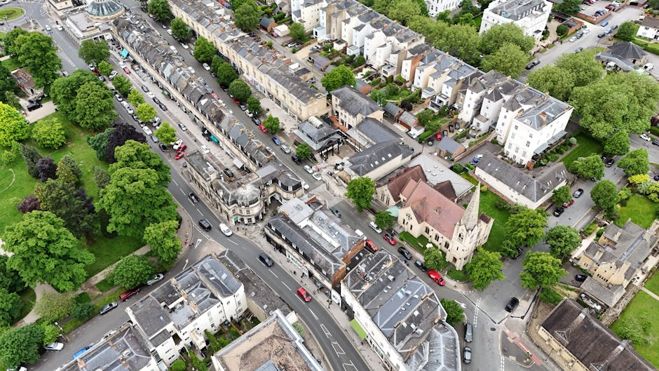 An aerial view of a residential street featuring a mix of terraced and detached houses, with a small church having a pointed steeple visible among buildings. The street has parked cars along both sides and a few vehicles in motion, with some parts of the road blocked for moving activities. Trees and green spaces line parts of the road and surrounding neighborhoods, providing a lush backdrop. Inside the property, a man and a van are engaged in home relocation activities, with furniture and cardboard boxes arranged nearby. The van is positioned at the curb, and some furniture, wrapped in protective materials such as blankets and plastic wrap, is being loaded or unloaded. The scene depicts the process of furniture transport and packing during a house removal, with the setting illuminated by natural daylight, emphasizing the urban environment and the logistics involved in moving services delivered by Man and Van Finsbury Park.