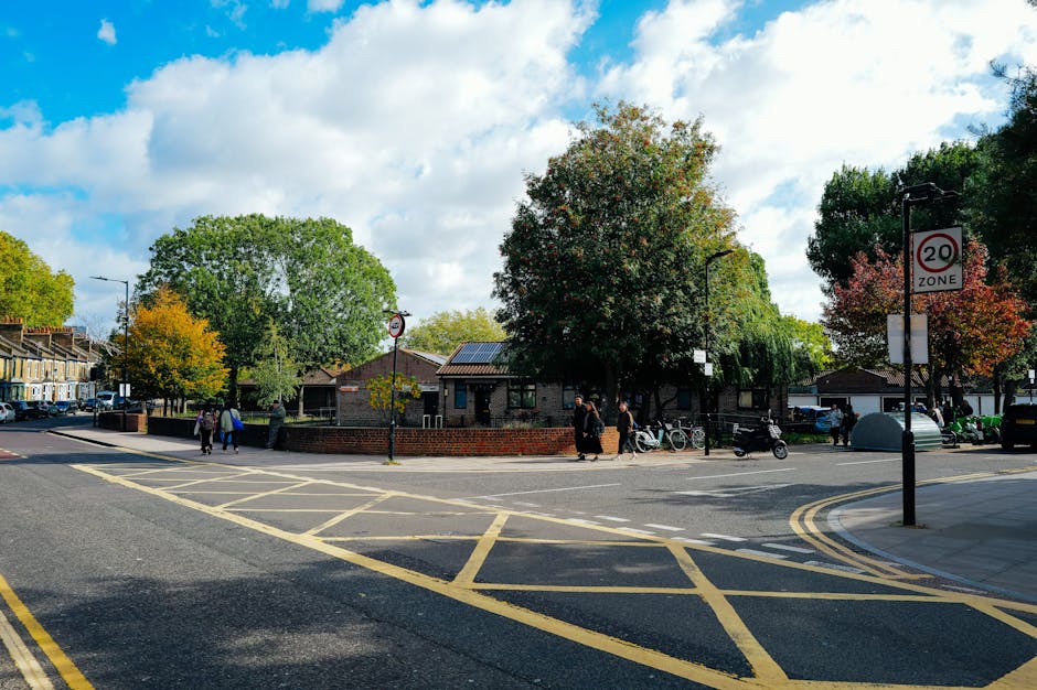 A street scene on Stroud Green Road in Finsbury Park showing a small public space with a brick-walled garden area surrounded by several mature trees with green and autumnal foliage, and a few pedestrians walking along the sidewalk. In the background, there are residential buildings, utility poles, and parked cars. The foreground features a road with yellow box markings, a 20 mph speed limit sign, and a lamppost, all under a partly cloudy sky. This setting illustrates an urban environment suitable for house removals and furniture transport activities, as part of professional home relocation services offered by Man and Van Finsbury Park.