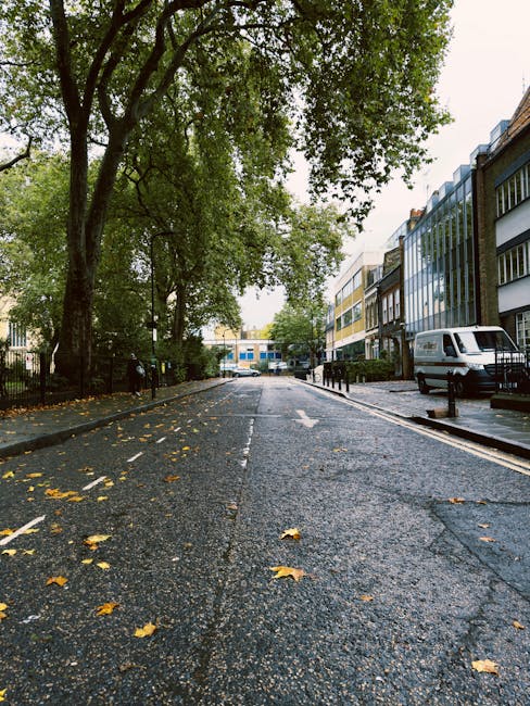 An aerial view of a residential street featuring a mix of terraced and detached houses, with a small church having a pointed steeple visible among buildings. The street has parked cars along both sides and a few vehicles in motion, with some parts of the road blocked for moving activities. Trees and green spaces line parts of the road and surrounding neighborhoods, providing a lush backdrop. Inside the property, a man and a van are engaged in home relocation activities, with furniture and cardboard boxes arranged nearby. The van is positioned at the curb, and some furniture, wrapped in protective materials such as blankets and plastic wrap, is being loaded or unloaded. The scene depicts the process of furniture transport and packing during a house removal, with the setting illuminated by natural daylight, emphasizing the urban environment and the logistics involved in moving services delivered by Man and Van Finsbury Park.